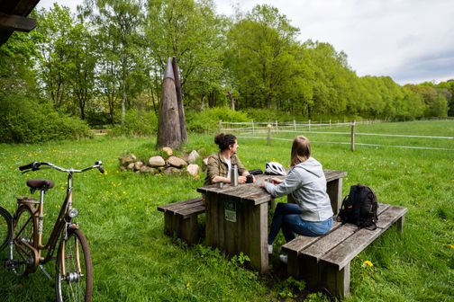 Zwei Radfahrer trinken auf einem malerischen Rastplatz am Ochsenweg bei Neumünster einen Kaffee, im Hintergrund ist eine Ochsen-Hörner-Skulptur zu sehen.